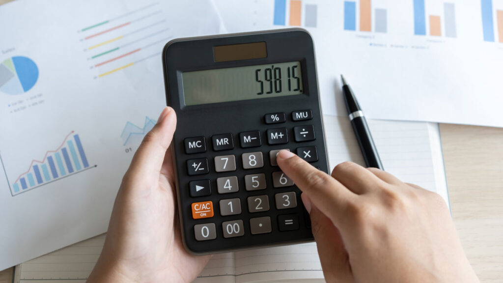 Hand, die einen Taschenrechner bedient, um die Gesamtkosten eines Mitarbeiters zu berechnen, im Hintergrund liegen Finanzdiagramme.